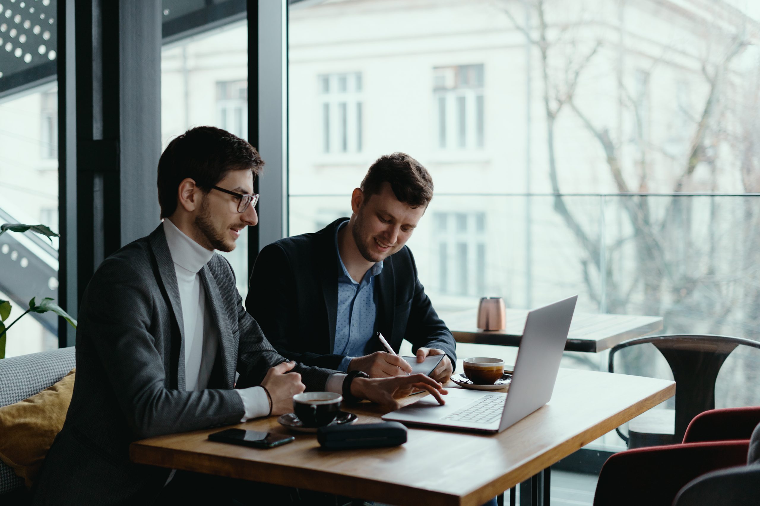 Two businessmen talking about new opportunities sitting with laptop at desk near large window, planning project, considering business offer, sharing ideas while drinking coffee together